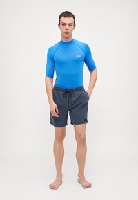 Blå kortermet rashguard og mørke shorts med snøring. Glatt tekstur, atletisk passform, med en logo på øvre venstre side.