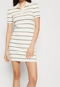 Femme portant une robe blanche à manches courtes en côtes, avec des rayures horizontales rouges, vertes et jaunes, et un petit logo vert sur la poitrine.