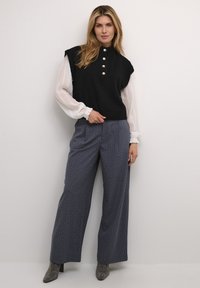 Black sleeveless sweater with pearl buttons, layered over a white blouse with ruffled sleeves. Navy wide-leg pants with small white dots.
