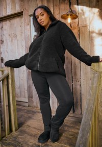 Musta fleece-takki, jossa on vetoketju edessä, ylisuurta mitoitusta, ja sivutaskut, yhdistettynä mustiin legginsseihin ja mustiin nilkkasaappaisiin puuporrasta pitkin.