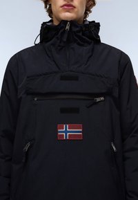 Mørkeblå jakke lavet af syntetisk stof, med halv-lynlås, brystlomme med velcrolåg, og et broderet norsk flag-patch.