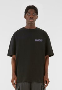 Tricou supradimensionat negru din bumbac, cu mâneci scurte și decolteu rotund. Text grafic notabil de culoare mov pe piept.