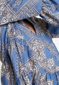 Modré šaty se vzorem, které mají jemné bílé a námořnické designy, výstřih do V a volné, vypasované rukávy vyrobené z lehkého materiálu.