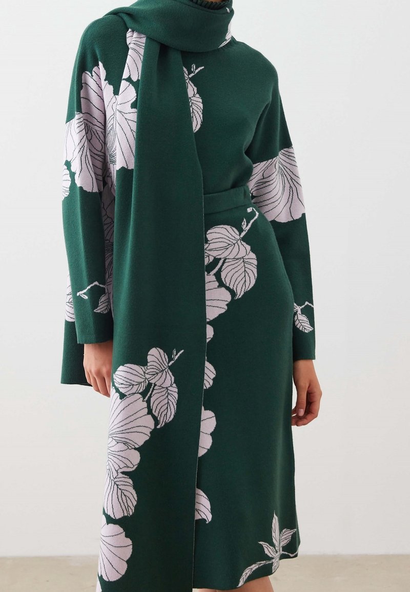 Robe en maille vert foncé et écharpe assortie ornées de grands motifs de feuilles rose pâle portées par une personne devant un fond uni.