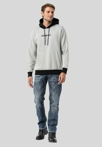Hellgrau Hoodie mit schwarzen Akzenten und Kordeln, Fronttasche, kombiniert mit blauen Jeans und schwarzen Schnürstiefeln. Modell steht vor grauem Hintergrund.
