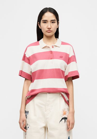 Polo rayé rose et crème, avec col, trois boutons et un logo. Associé à un pantalon crème orné de motifs brodés.