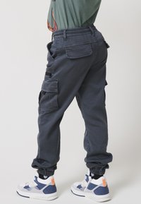 Temno sivi cargo hlače z elastičnim pasom, stranskimi žepi in zoženimi manšetami. Nosijo se z belimi in mornarsko modrimi superge, ki imajo oranžne poudarke.