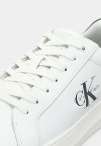 Witte Calvin Klein sneakers met witte veters, zwart CK-logo en branding op de zijkant, zichtbare stiksels en een dikke zool.