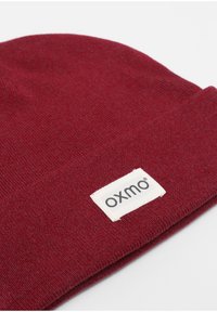 Burgundy knit beanie made of soft material, featuring a folded cuff with a white label displaying "oxmo." Smooth texture with no patterns.