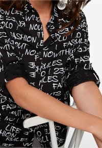 Chemise noire à bouton avec des motifs de texte blancs incluant "FASHION," "NO RULES," et "LIFE." Le tissu a une texture lisse. Manches retroussées.