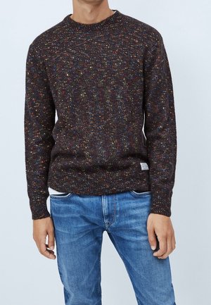 Mann trägt einen dunkel multicolor gesprenkelten Strickpullover und blaue Jeans, steht vor einem schlichten hellen Hintergrund.