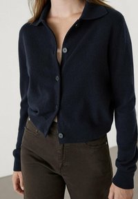 Granatowy wełniany kardigan z kołnierzem, z guzikami z przodu i prążkowanymi mankietami, w połączeniu z ciemnobrązowymi spodniami. Miękka tekstura i dopasowany fason.