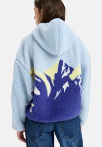 Lys blå, fluffy, hooded jakke med et lilla og gult bjerggrafik på ryggen. Blød tekstur med en afslappet pasform.