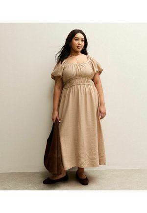 Vrouw met donker haar staat, draagt een beige lange jurk, houdt een bruine tas vast en draagt zwarte platte schoenen tegen een eenvoudige lichte muur.