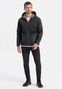 Giubbotto di jeans nero con cappuccio grigio, tasche frontali e chiusura a zip. Pantaloni neri slim-fit e scarpe alte nere completano l'outfit.