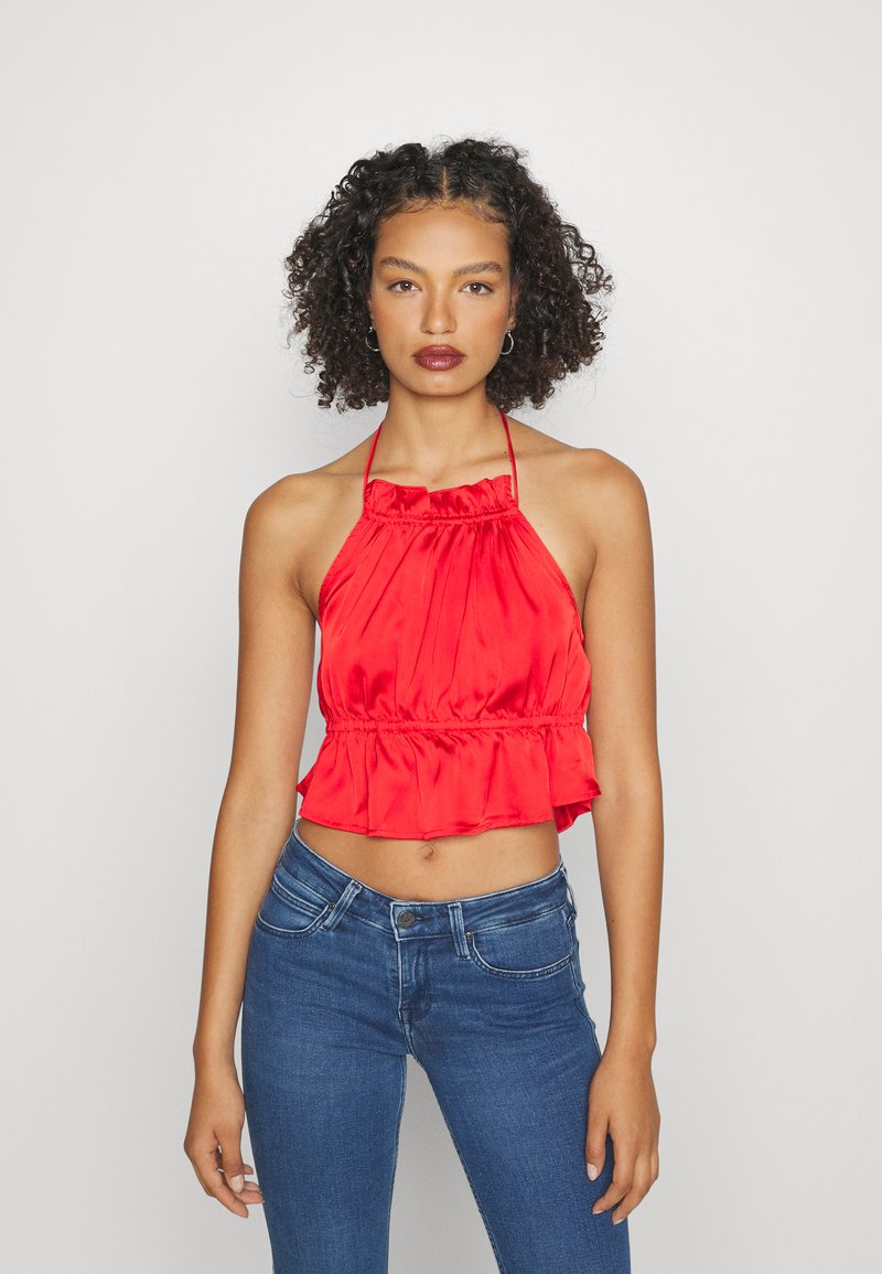 Top halter em cetim vermelho com decote em babados e barra peplum. Textura suave, comprimento cropped e laços ajustáveis nas costas. Combinado com jeans azuis.