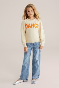 Krämfärgad tröja med orange "DANCE" text, kombinerad med ljusblå vida jeans prydda med silverdetaljer, och metallic silverfärgade lägenheter.