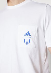 Hvitt bomull t-skjorte med brystlomme. Har blå logoer, inkludert en adidas-emblem og en krigersk grafikk nedenfor.