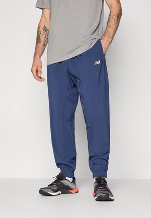 Pantalon de sport bleu marine avec des poignets élastiques et des poches, associé à des chaussures de course noires avec des accents rouges et le logo "MINIMUS".