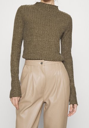 Kobieta w oliwkowo zielonym swetrze prążkowanym z długimi rękawami i beżowych, wysokim stanem, plisowanych skórzanych spodniach na białym tle.