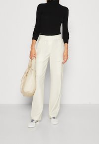 Pantaloni bianchi a gamba larga con tasche cargo laterali, abbinati a un dolcevita nero a coste. Gli accessori includono una borsa beige e scarpe da ginnastica bianche.