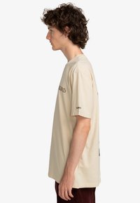 Beige katoenen T-shirt met korte mouwen, voorzien van een logo aan de voorkant in bruin en een grafische print op de achterkant. Ontspannen pasvorm, zachte textuur en rechte snit.