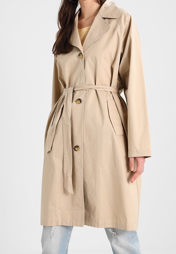 Femme portant un trench-coat beige ceinturé avec des boutons, un large col et des poches latérales, par-dessus un jean bleu clair déchiré.