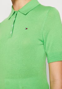 Zelená pletená polo tričko s límečkem, krátkými rukávy a třemi knoflíky. Na hrudi se nachází malé logo v červené, bílé a modré barvě.