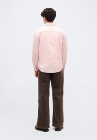 Homme de dos portant une chemise à manches longues rose clair, un pantalon large marron foncé et des chaussures noires sur un fond uni.