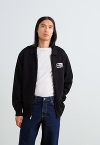 Zwarte zip-up jas met een puntige kraag en geborduurd logo op de linker borst, gecombineerd met een effen wit shirt en donkerblauwe jeans.