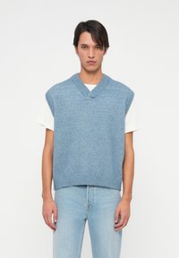 Jovem vestindo um colete de malha azul claro sem mangas sobre uma camiseta branca e jeans azul claro, contra um fundo branco simples.
