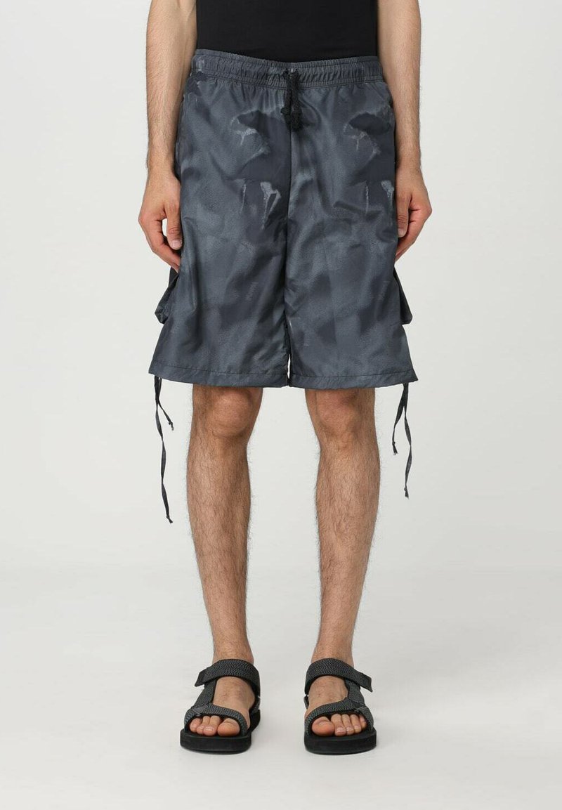 Pantaloni corti grigi di lunghezza media realizzati in un tessuto leggero con un motivo sottile, caratterizzati da una vita con cordoncino e tasche laterali. Sono indossate delle ciabatte piatte.