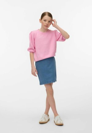 Mujer joven de pie, tocándose el pelo con la mano, vestida con un suéter rosa, falda vaquera azul y zapatillas blancas sobre fondo blanco.