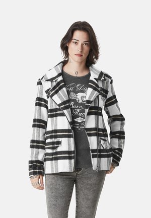 Personne portant une veste zippée à carreaux noirs, blancs et gris par-dessus un t-shirt graphique foncé et un jean gris, debout devant un fond blanc.