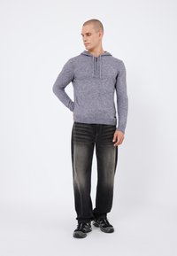 Grau gestrickter Kapuzenpullover mit Kordelzug, kombiniert mit schwarzen verwaschenen Jeans und schwarzen Sportschuhen mit grünen Akzenten. Mann steht in neutraler Pose.
