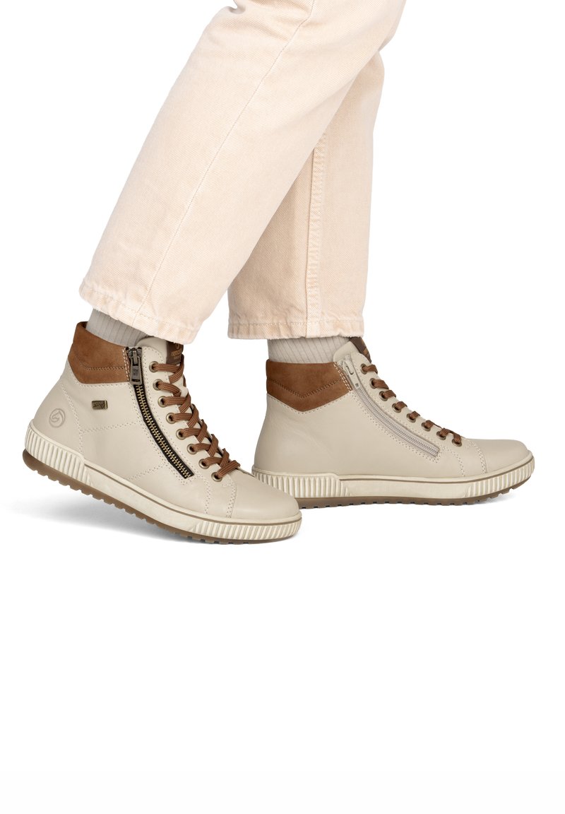 Personne portant des baskets montantes beiges avec des accents marron, des fermetures éclair latérales et des lacets assortis à un pantalon clair.