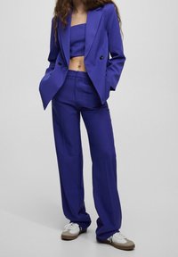 Ensemble deux pièces en violet vif, comprenant un haut court et un blazer ajusté avec boutons, associé à un pantalon ample et des baskets.
