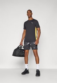 Schwarz sportliches T-Shirt mit gelbem Seitenstreifen, bedruckte schwarze Shorts, schwarze Turnschuhe, weiße Socken und eine schwarze Sporttasche. Einfaches Design mit Logo-Akzenten.