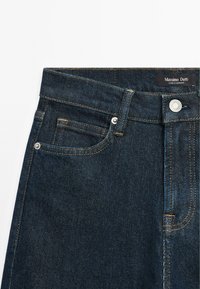Jeans in denim blu scuro con taglio dritto, dotati di una tasca frontale, cuciture evidenti e un'etichetta con il marchio sulla vita.