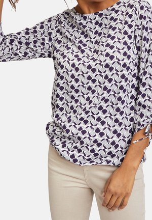 Weiße Bluse mit einem lila geometrischen Muster, Rundhalsausschnitt und Dreiviertelärmeln mit Bindebändern an den Bündchen. Weicher Stoff.