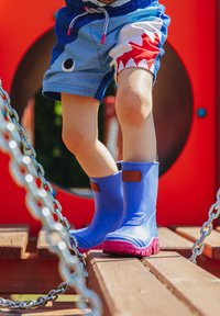 Blaue Gummistiefel mit pinken Sohlen und braunen Lederakzenten, getragen auf einer Holzoberfläche, Teil eines Spielplatzes.