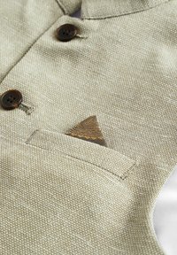 Jasnobeżowa bezrękawnik z teksturowanej tkaniny, brązowe guziki oraz wzorzysta poszetka wystająca z kieszeni na piersi.