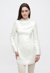 Korte, aansluitende jurk van gestructureerde witte stof met lange mouwen en een hoge halslijn. Heeft een rechte silhouet en minimale details.