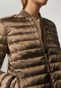 Bruine pufferjack met een gekwalteerd ontwerp, ribbelkraag en ritssluiting. Heeft een gladde, glanzende textuur en een aangesloten silhoutte.