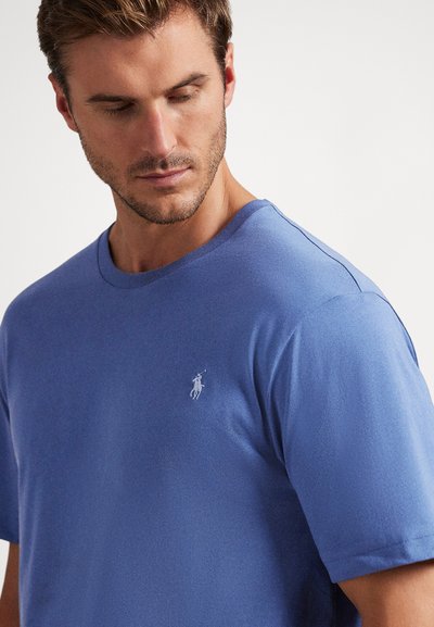Vīriešu īsroku zilā T-krekla, izgatavots no mīksta auduma, ar apaļu izgriezumu un mazu baltu izšūtu logotipu kreisajā krūtī.