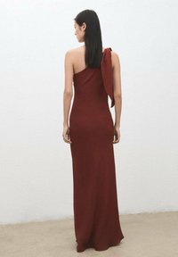 Asymetryczna bordowa sukienka o miękkiej fakturze, z kokardą na ramieniu i dopasowanym krojem sięgającym do kostek.