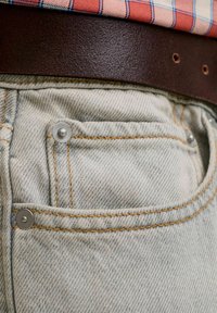Poche de jean en denim gris clair avec surpiqûres orange, rivets argentés, portée avec une ceinture en cuir marron foncé et une chemise à carreaux rouge.