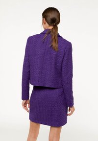 Woman with brown ponytail wearing a textured purple jacket and matching skirt, standing facing away against a plain white background.