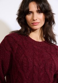 Bordowy sweter z dzianiny z grubym wzorem warkocza, okrągłym dekoltem i fakturowaną tkaniną. Zbliżenie na szczegóły ubrania.