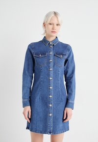 Τζιν φόρεμα με μακριά μανίκια, κουμπιά μπροστά, κολάρο, δύο τσέπες στο στήθος και ανοιχτό μπλε χρώμα. Διαθέτει ίσια γραμμή και ελαφριά κλίση στο τελείωμα.